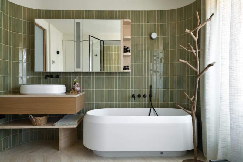 La salle de bains ramenée à la lumière naturelle, renoue avec les courbes, par son parement en céramique et sa baignoire (Agape). Au sol, le parquet fait place à des lames de travertin