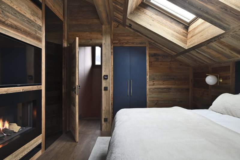 La master suite tutoie le ciel, réchauffée par une cheminée au gaz. L’agencement est ici totalement absorbé par le vieux bois hormis ce placard confectionné en textile, Dedar